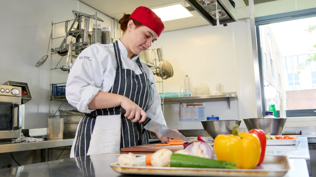 Photo of a student using a chopping knife and board to prepare vegetables in a kitchen